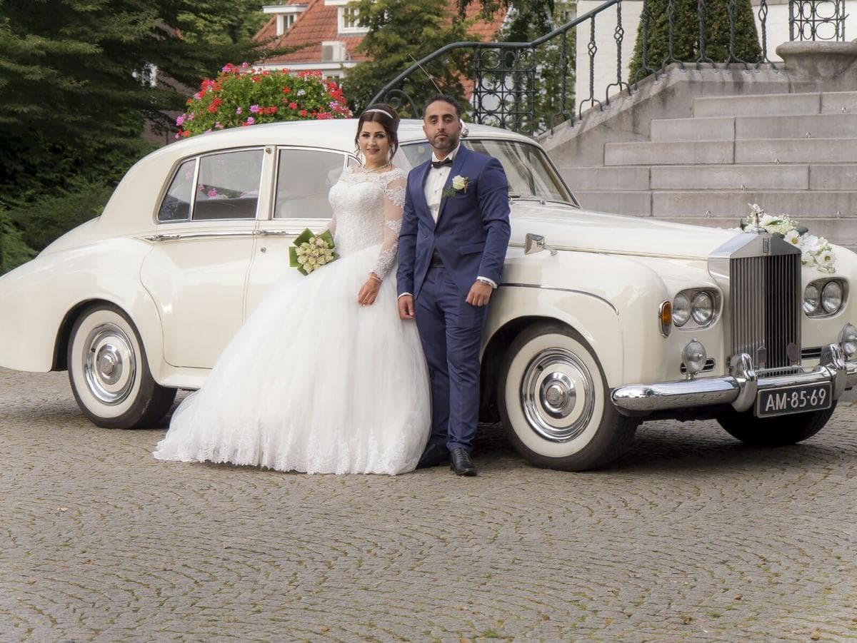 Bruid in witte jurk en bruidegom in blauw pak poseren voor een witte klassieke trouwauto